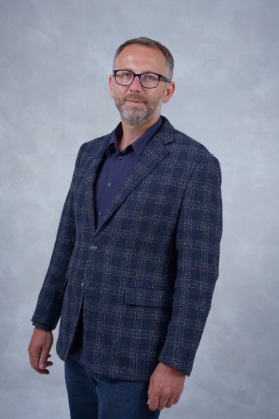 Mężczyzna w okularach i niebieskiej marynarce w kratę, na rozmytym szarym tle. Częściowy widok sylwetki, skierowany lekko w lewo.