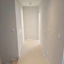 Minimalistyczny korytarz w jasnych, neutralnych barwach; widoczne ukryte drzwi, gładkie ściany, jednolite podłogi i nowoczesne oświetlenie punktowe.