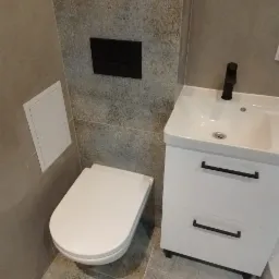 Nowoczesna łazienka z podwieszaną toaletą, białą szafką umywalkową z czarną armaturą i szarymi płytkami z delikatnym wzorem.