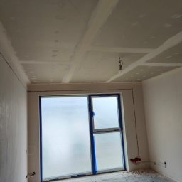 Palian Professional - Pokój w trakcie prac wykończeniowych: zabezpieczone okno folią, ściany i sufit po gładziowaniu, widoczne łączenia płyt gipsowych. Podłoga zakryta folią ochronną.