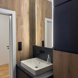 Palian Professional - Nowoczesna łazienka z drewnopodobnymi płytkami, czarnym blatem i armaturą. Lustro z podświetleniem LED. Widoczny fragment drzwi i podłogi w innym pomieszczeniu.