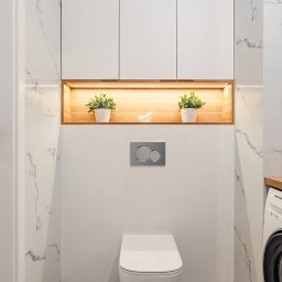 Palian Professional - Nowoczesna łazienka z podwieszaną toaletą, marmurowymi płytkami i oświetloną wnęką z roślinami. Minimalistyczny design, pralka z blatem po prawej stronie.