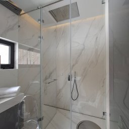 Palian Professional - Nowoczesna łazienka z kabiną prysznicową typu walk-in, wykończona białym marmurem. Widoczny transparentny fotel i okno z widokiem na drzewa.