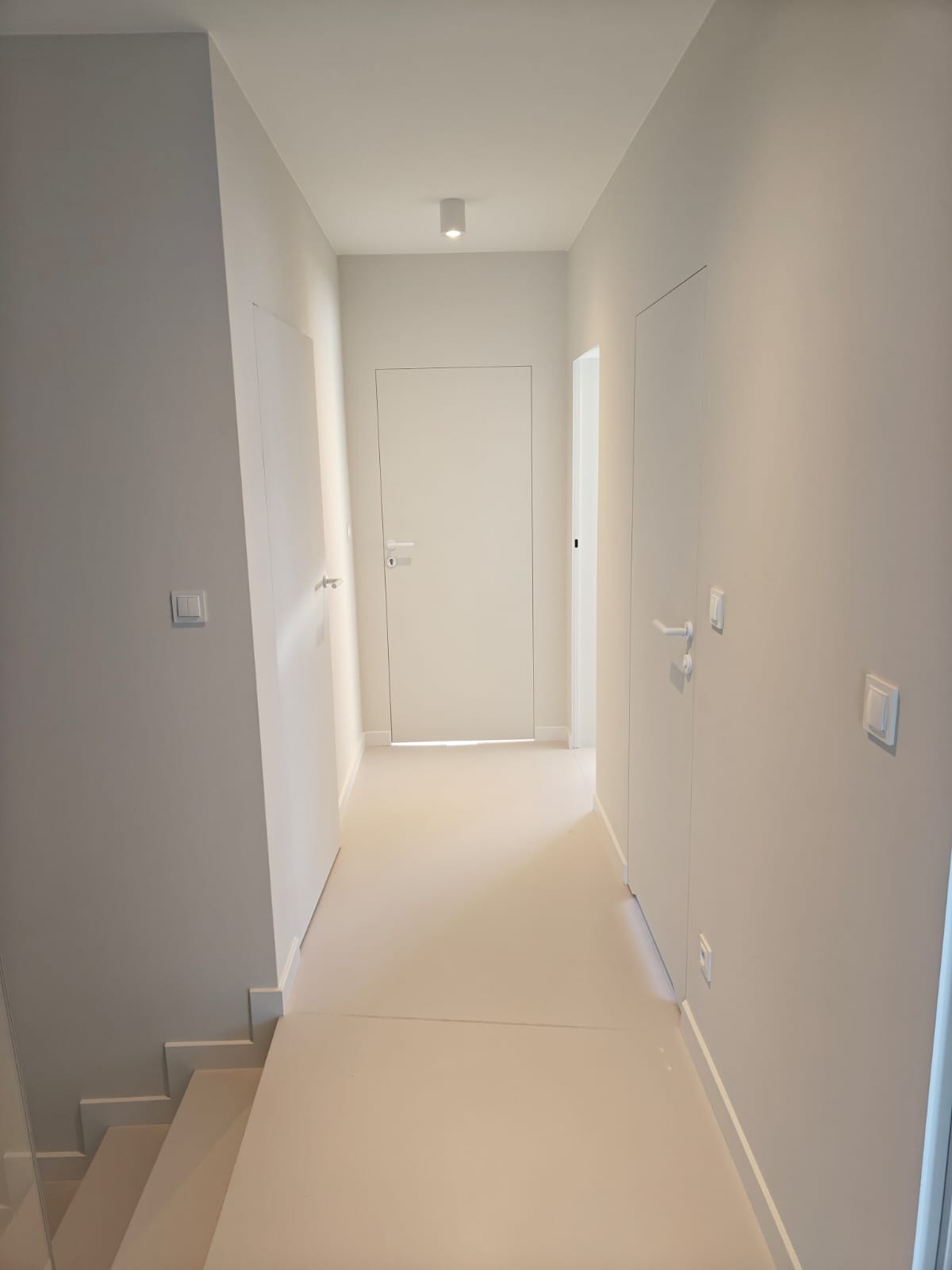Minimalistyczny korytarz w jasnych, neutralnych barwach; widoczne ukryte drzwi, gładkie ściany, jednolite podłogi i nowoczesne oświetlenie punktowe.