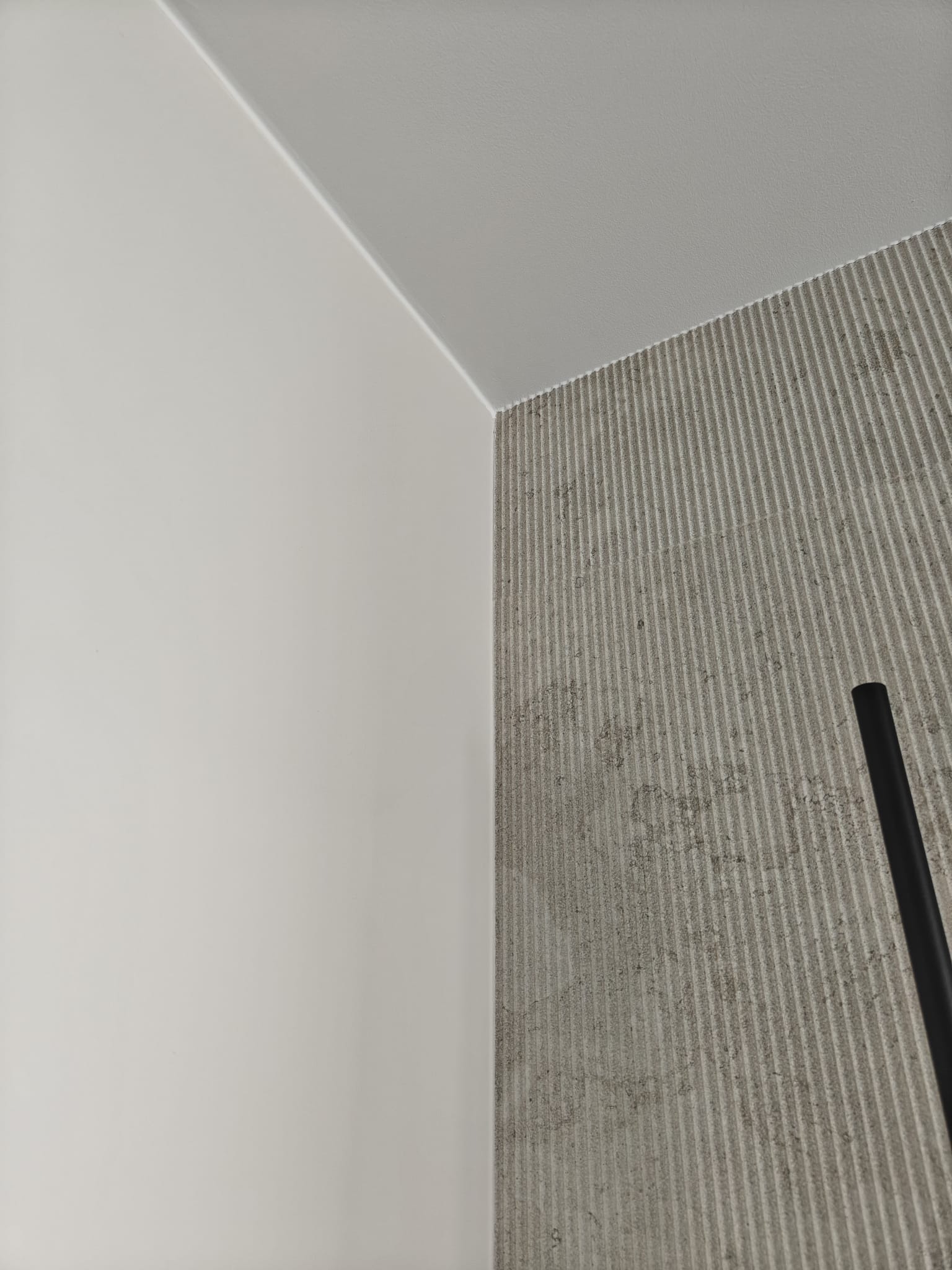 Fragment narożnika pokoju z gładką, pomalowaną na biało ścianą, białym sufitem i ścianą pokrytą szarą, ryflowaną okładziną imitującą beton, z fragmentem czarnego elementu (np. rury) po prawej stronie.