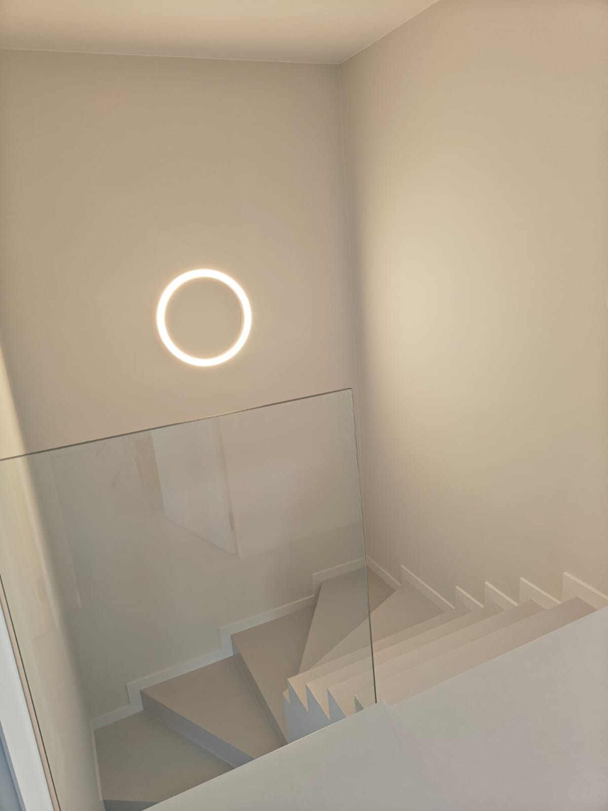 Nowoczesne, minimalistyczne schody w odcieniach bieli w Warszawie, z szklaną balustradą i okrągłym, podświetlanym plafonem na ścianie.