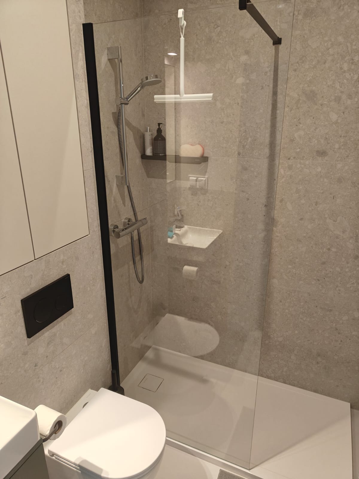 Nowoczesna łazienka z kabiną prysznicową typu walk-in z czarną ramą, umywalką nablatową i toaletą z czarnym przyciskiem spłukiwania, wykończona płytkami imitującymi kamień.