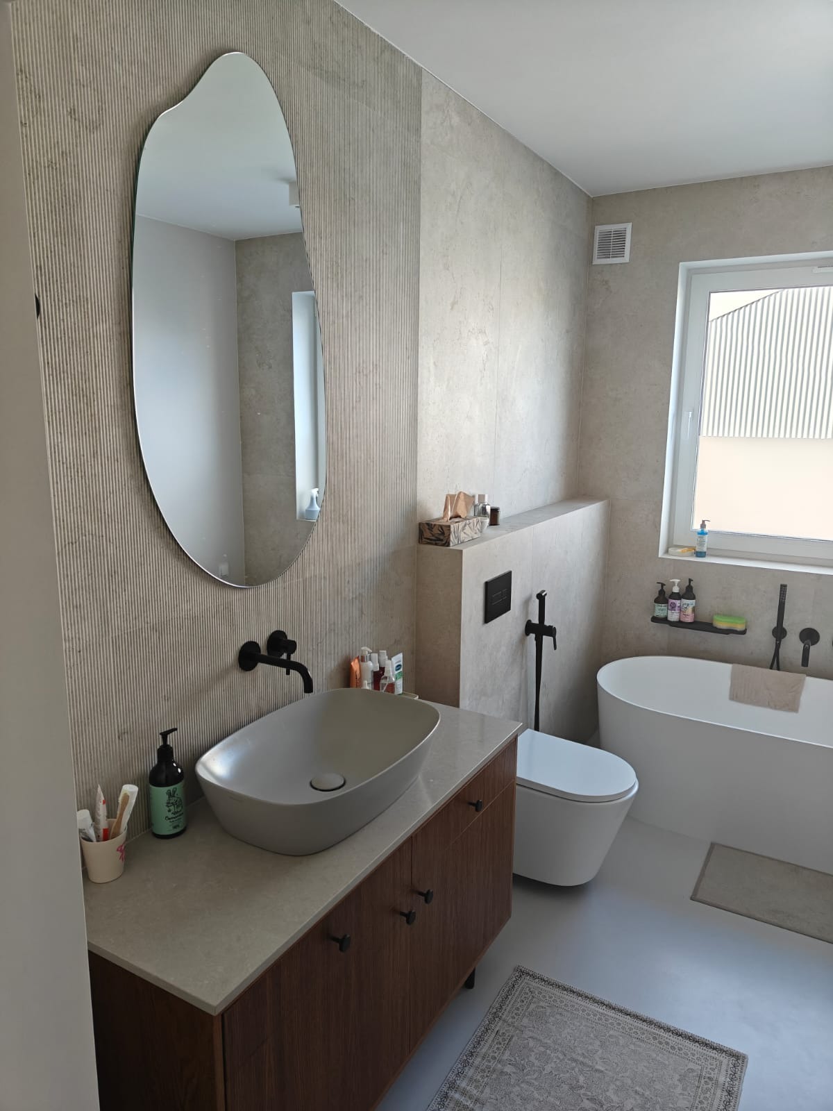 Nowoczesna łazienka z owalnym lustrem, umywalką nablatową, drewnianą szafką pod umywalką, beżowymi płytkami na ścianie i wolnostojącą wanną.