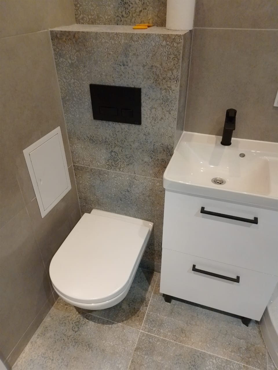 Nowoczesna łazienka z podwieszaną toaletą, białą szafką umywalkową z czarną armaturą i szarymi płytkami z delikatnym wzorem.