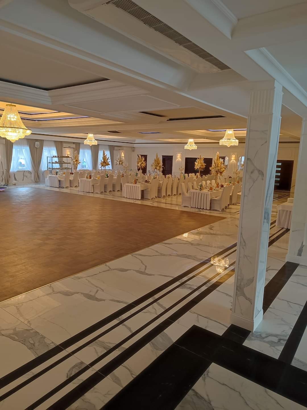 Elegancka sala balowa z parkietem, marmurową posadzką i kryształowymi żyrandolami. Stoły nakryte białymi obrusami, gotowe na przyjęcie. Widok z perspektywy podłogi.