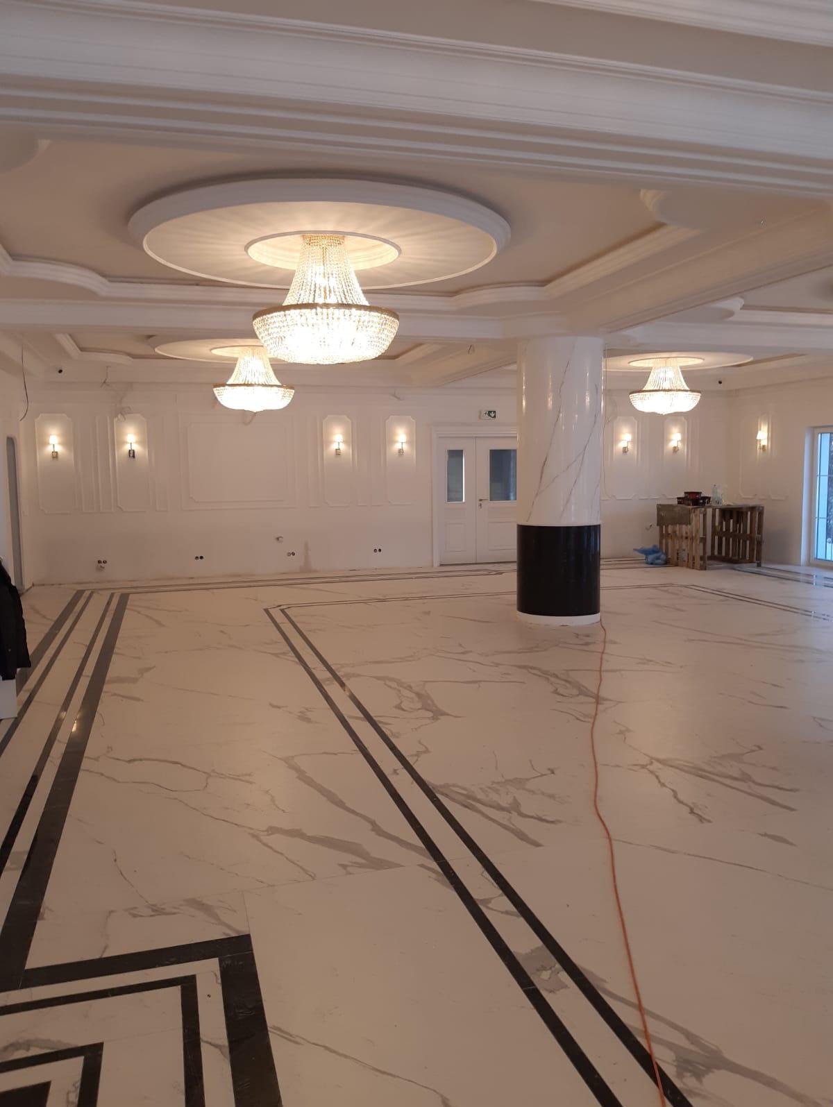 Eleganckie wnętrze po wykończeniu: marmurowa podłoga z czarnymi akcentami, kolumna w marmurze, ozdobne listwy ścienne i kryształowe żyrandole. Widok na przestronne, jasne pomieszczenie.