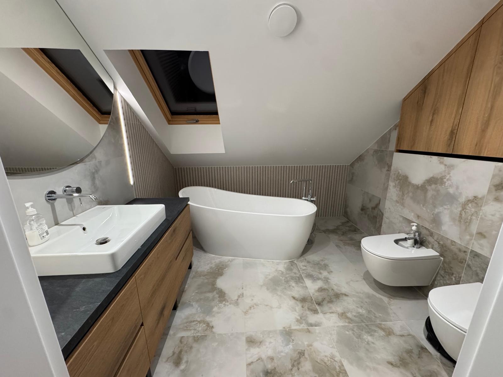 Nowoczesna łazienka na poddaszu z wolnostojącą wanną, umywalką nablatową na drewnianej szafce i marmurowymi płytkami. Okno dachowe doświetla wnętrze.