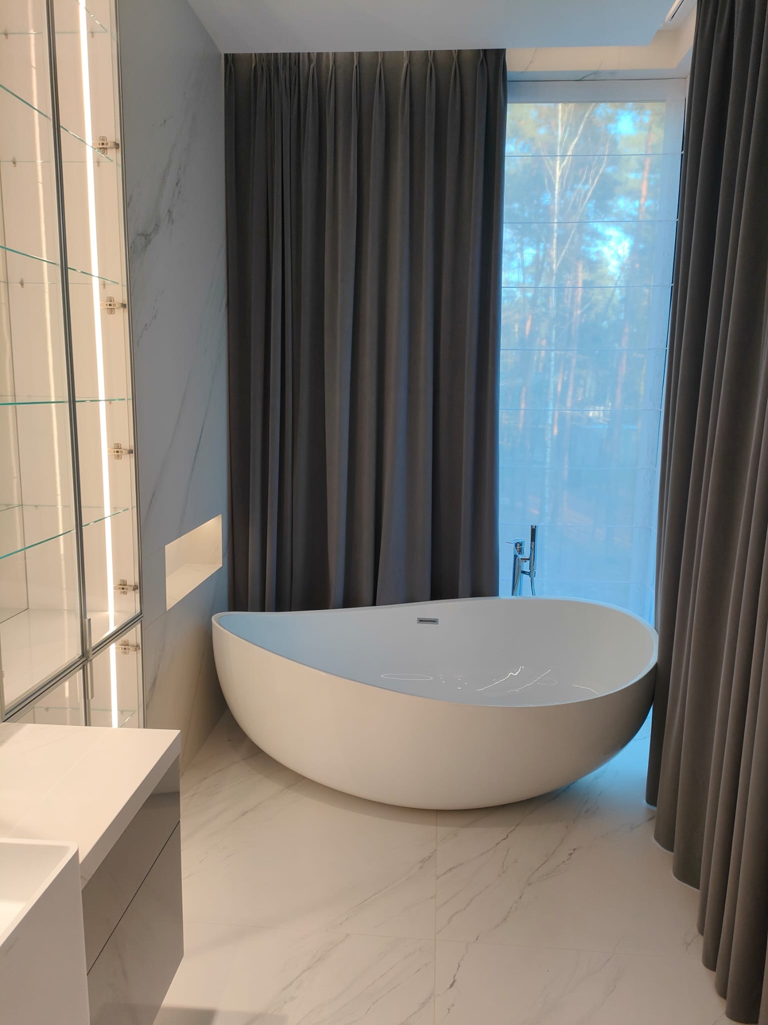 Nowoczesna łazienka z wolnostojącą wanną o organicznym kształcie, marmurową okładziną ścian i podłogi oraz szarymi zasłonami. Minimalistyczny design z oświetleniem LED.