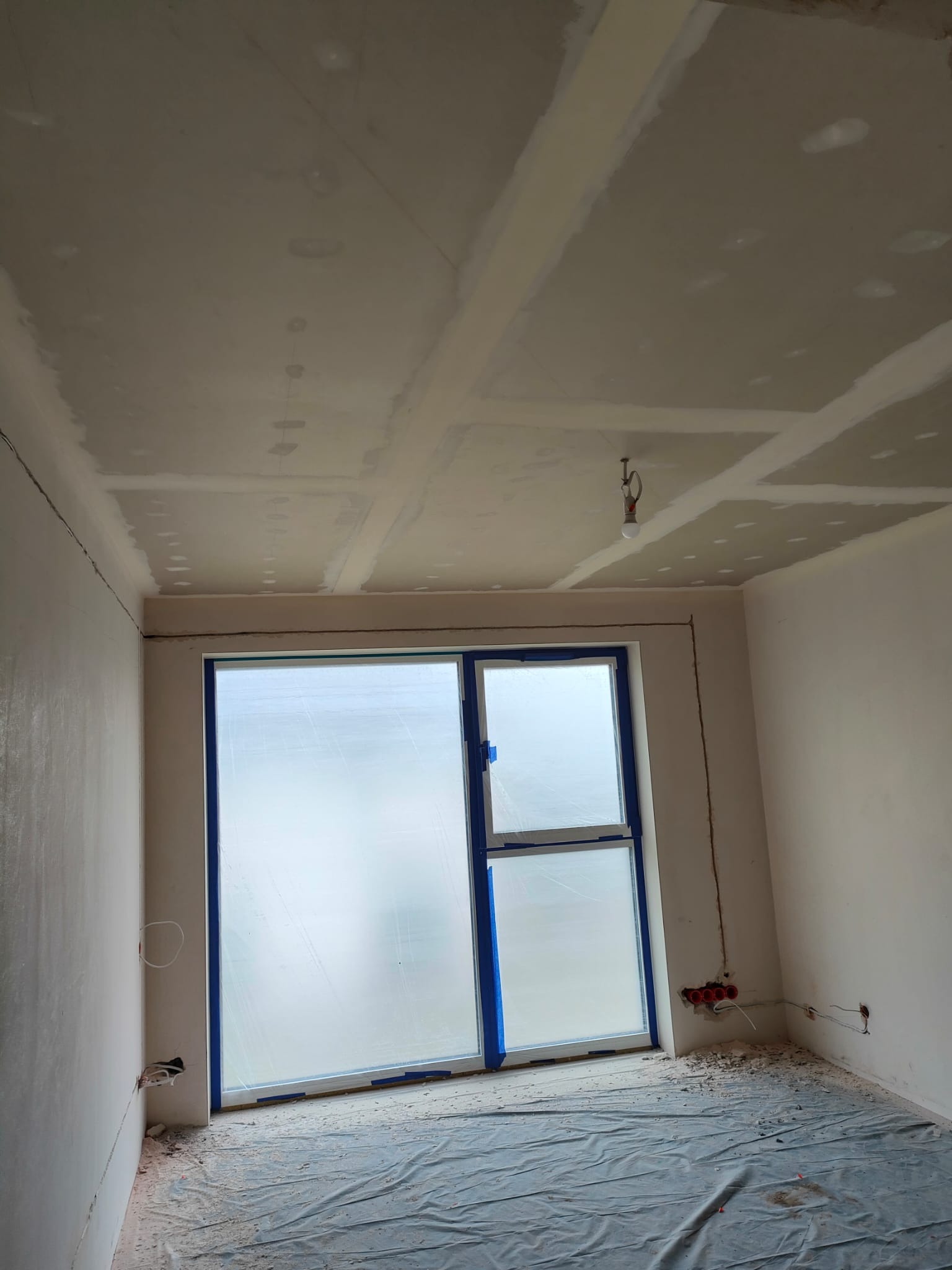 Pokój w trakcie prac wykończeniowych: zabezpieczone okno folią, ściany i sufit po gładziowaniu, widoczne łączenia płyt gipsowych. Podłoga zakryta folią ochronną.