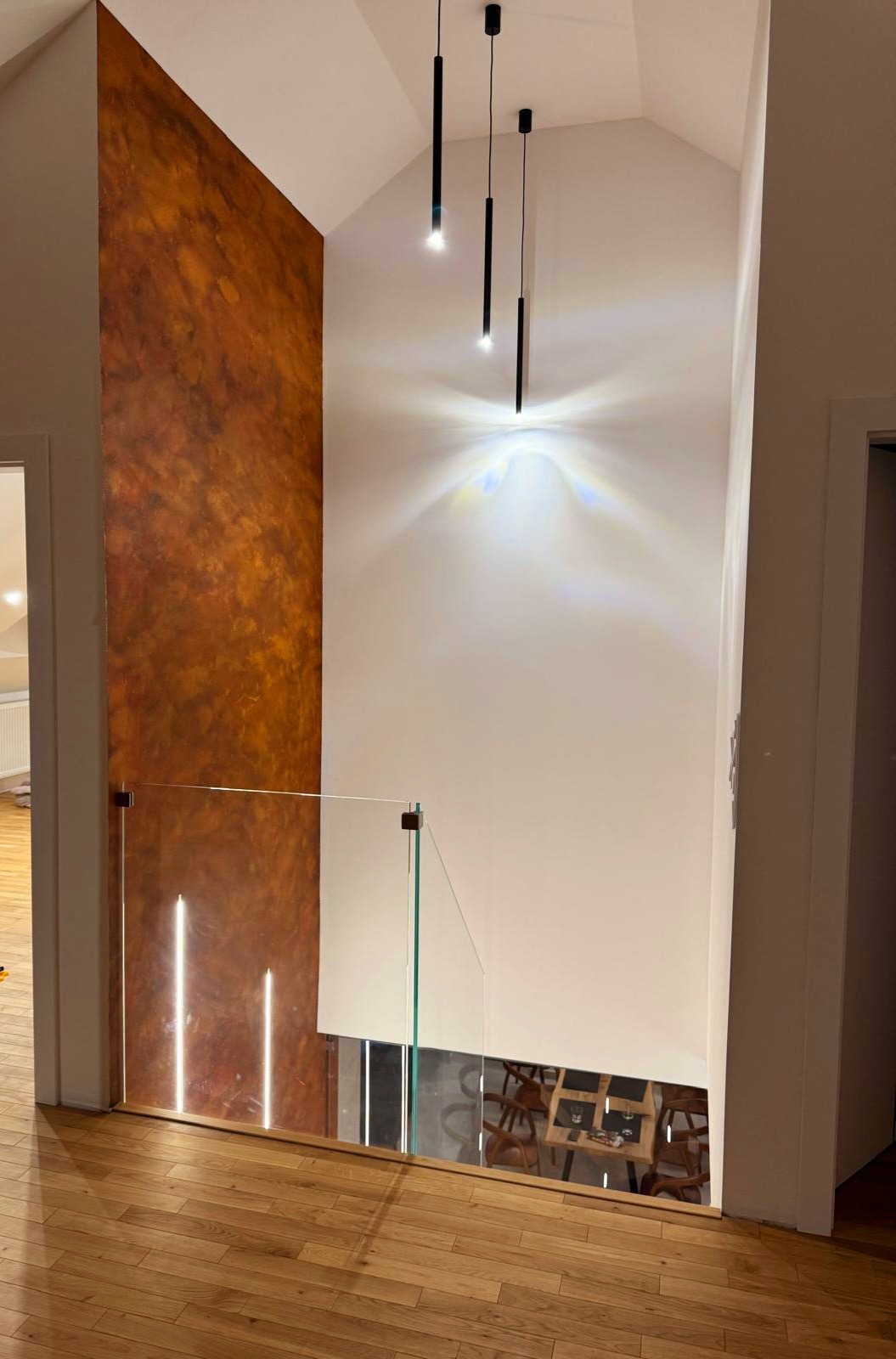 Nowoczesne wykończenie wnętrza: ściana z efektem rdzy, szklana balustrada i minimalistyczne lampy wiszące w holu z drewnianą podłogą. Detal aranżacji.