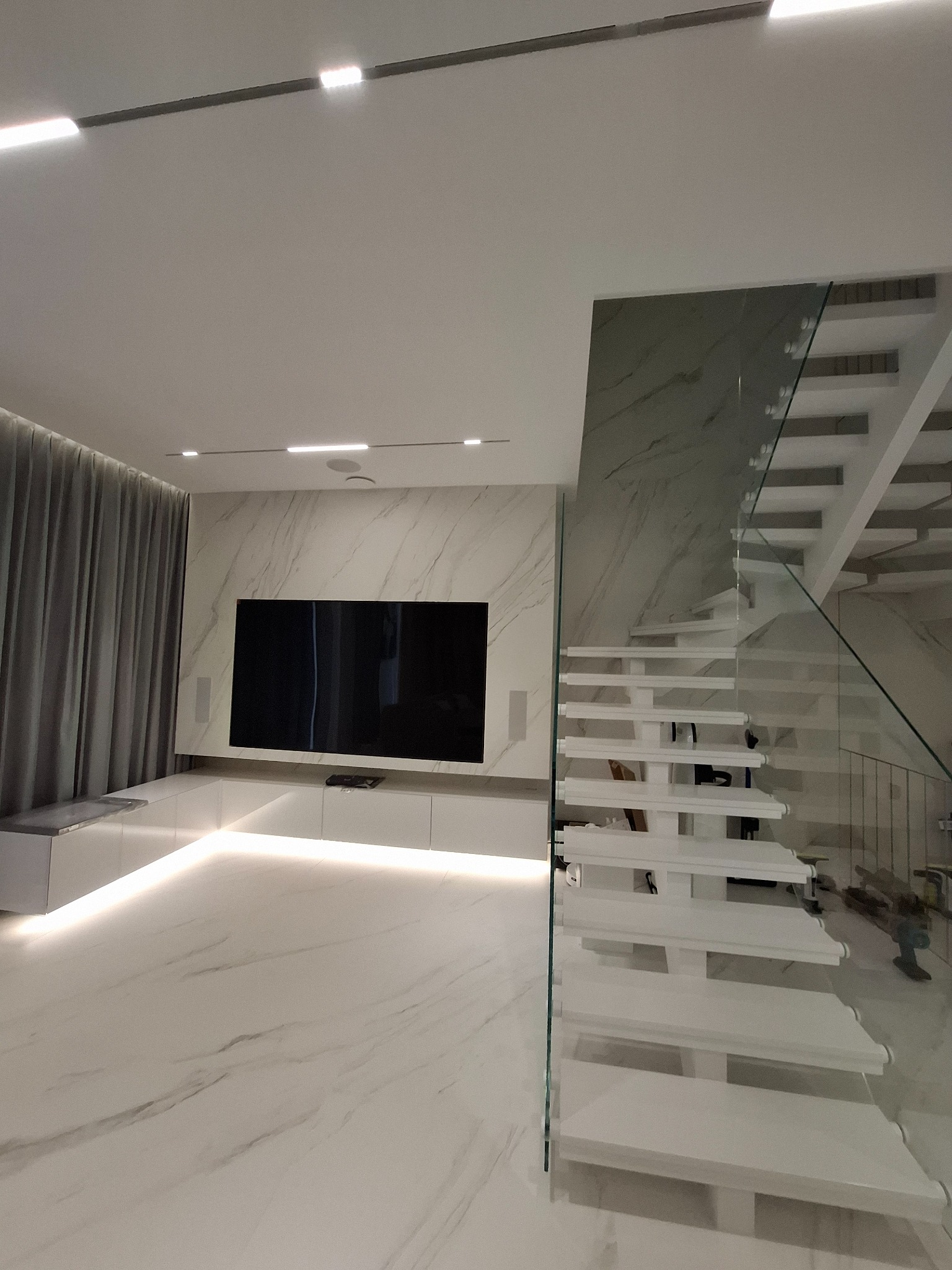 Nowoczesne wnętrze: białe marmurowe podłogi i ściany, minimalistyczne schody ze szklaną balustradą, podświetlana szafka RTV z czarnym telewizorem.