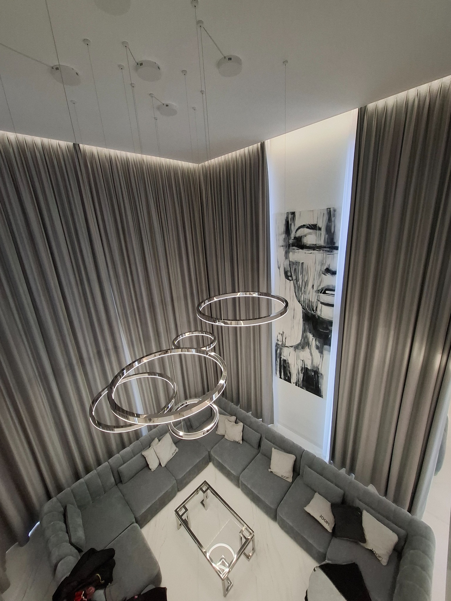 Elegancki salon z szarym narożnikiem modułowym, szklanym stolikiem i nowoczesną lampą pierścieniową. Na ścianie abstrakcyjny obraz w odcieniach szarości. Całość dopełniają długie, szare zasłony.