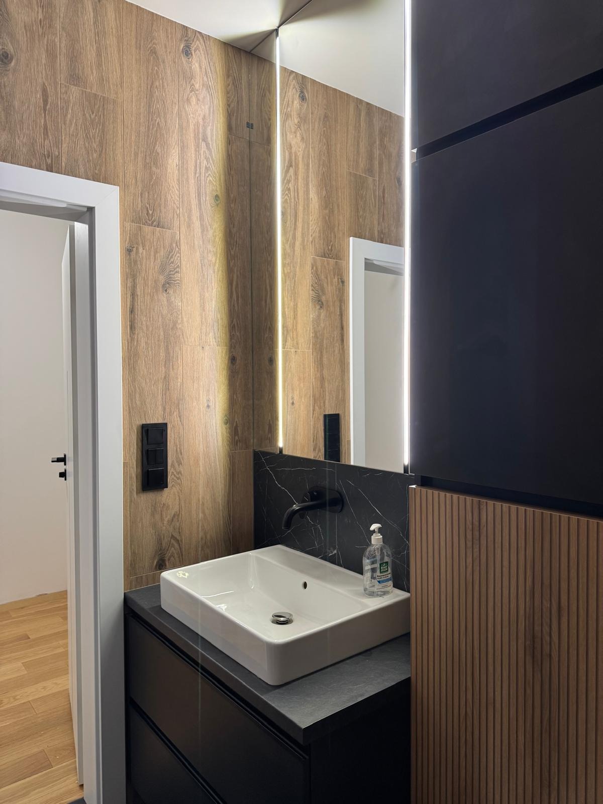 Nowoczesna łazienka z drewnopodobnymi płytkami, czarnym blatem i armaturą. Lustro z podświetleniem LED. Widoczny fragment drzwi i podłogi w innym pomieszczeniu.