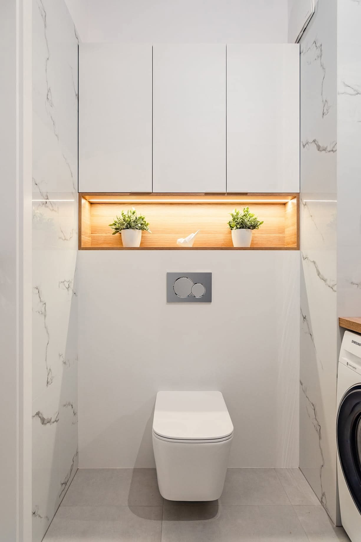Nowoczesna łazienka z podwieszaną toaletą, marmurowymi płytkami i oświetloną wnęką z roślinami. Minimalistyczny design, pralka z blatem po prawej stronie.