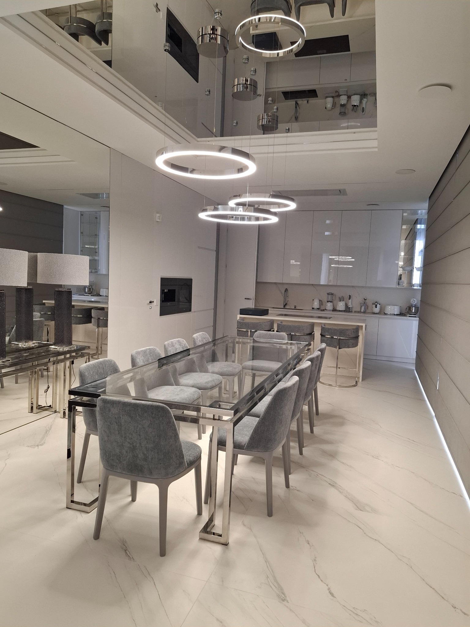 Nowoczesna jadalnia z marmurową podłogą, szklanym stołem i szarymi krzesłami. Lustrzany sufit odbija okrągłe lampy. Widoczna kuchnia w tle.