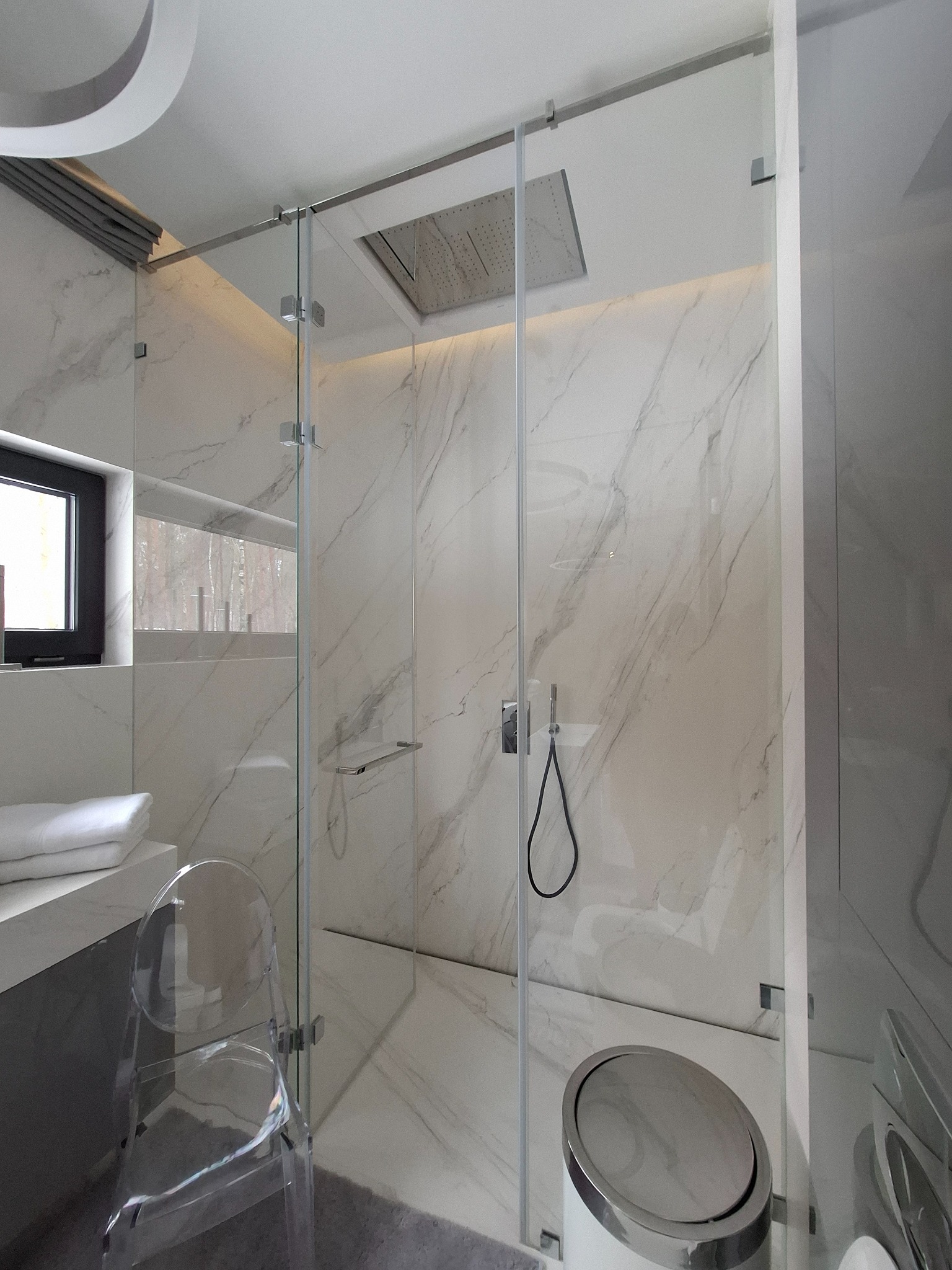 Nowoczesna łazienka z kabiną prysznicową typu walk-in, wykończona białym marmurem. Widoczny transparentny fotel i okno z widokiem na drzewa.