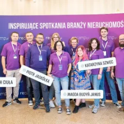 Grupa os&oacute;b w fioletowych koszulkach pozuje na tle baneru z napisem 'Inspirujące spotkania branży nieruchomości', trzymając tabliczki z imionami i nazwiskami oraz nazwami kont w mediach...