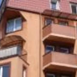 Fragment elewacji budynku wielorodzinnego z balkonami, widoczny dach z czerwonej dachówki ceramicznej, jasne okna PCV, elewacja w odcieniach pomarańczy, ujęcie z dołu.