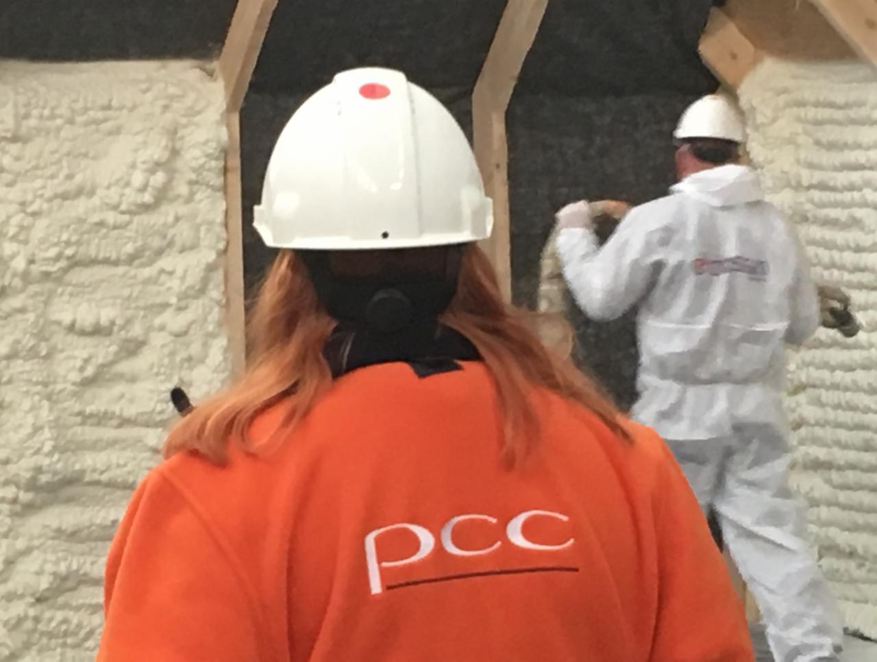 Pracownicy w kombinezonach ochronnych aplikują izolację natryskową wewnątrz budynku; widoczna struktura piany i logotyp firmy na pomarańczowej bluzie. Jeden z pracowników ma na głowie biały kask...