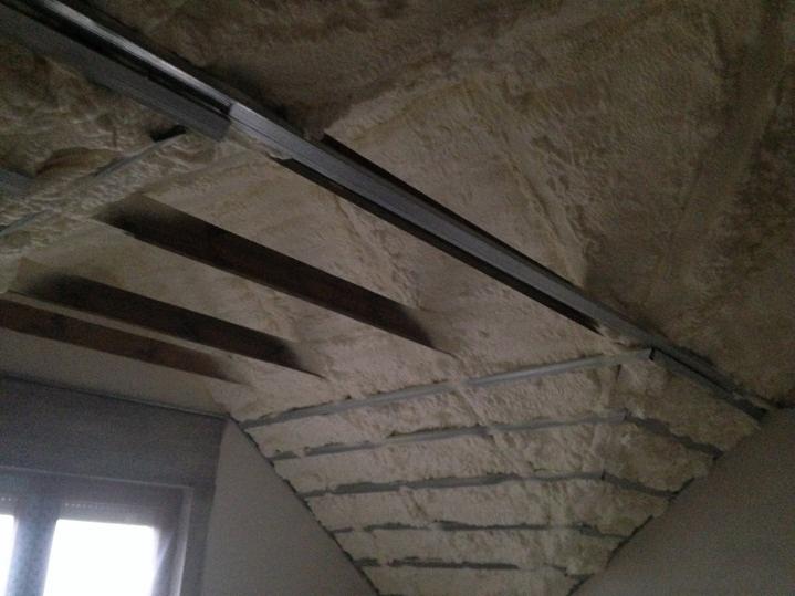 Poddasze z widoczną izolacją natryskową między krokwiami i profilami metalowymi, w tle okno.