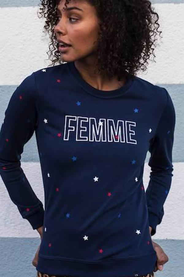 Kobieta w granatowej bluzie z długim rękawem, z białym napisem 'FEMME' i wzorem z czerwonych, białych i niebieskich gwiazdek, pozuje na tle szarej ściany.