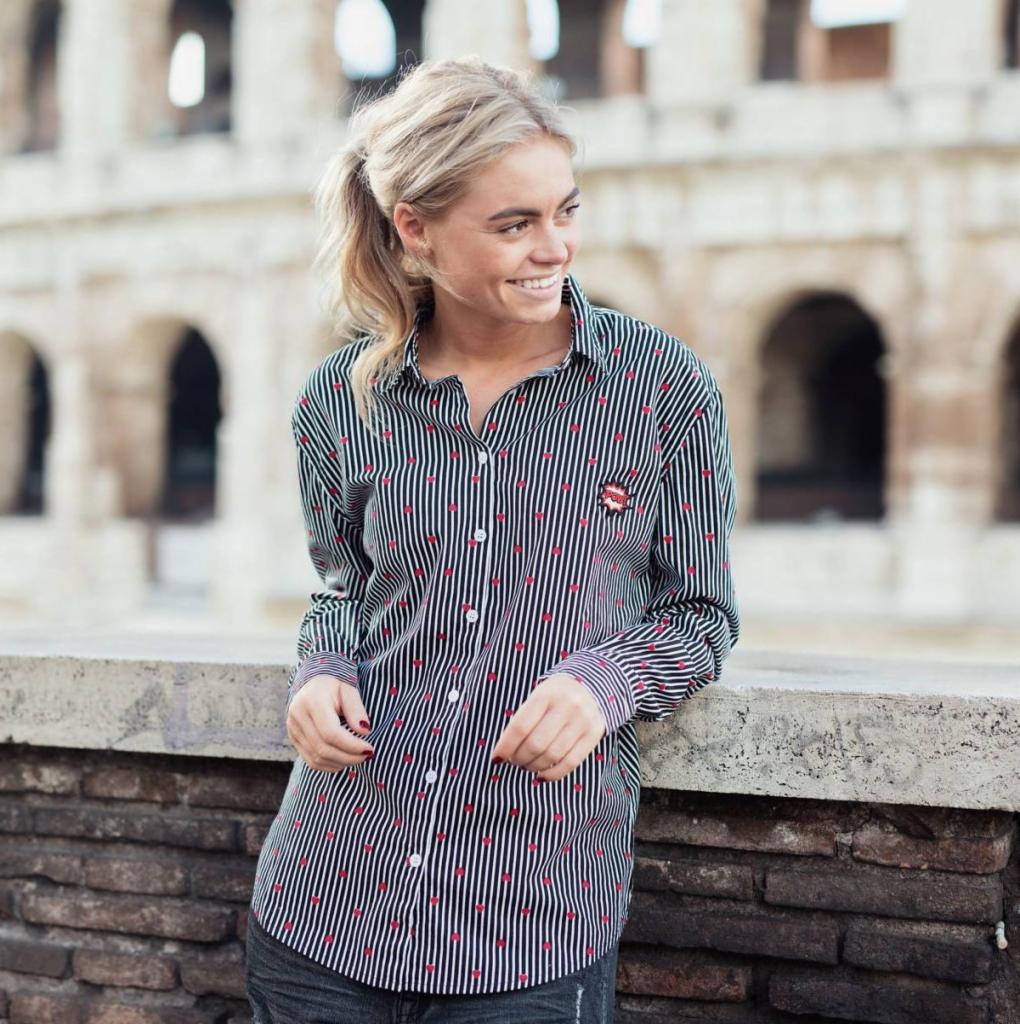 Uśmiechnięta blondynka w koszuli w paski z czerwonymi serduszkami, oparta o kamienny mur na tle Koloseum.