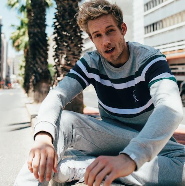 Młody mężczyzna w szaro-granatowej bluzie w paski i dresowych spodniach, siedzący na murku w miejskim otoczeniu z palmami i budynkami w tle.