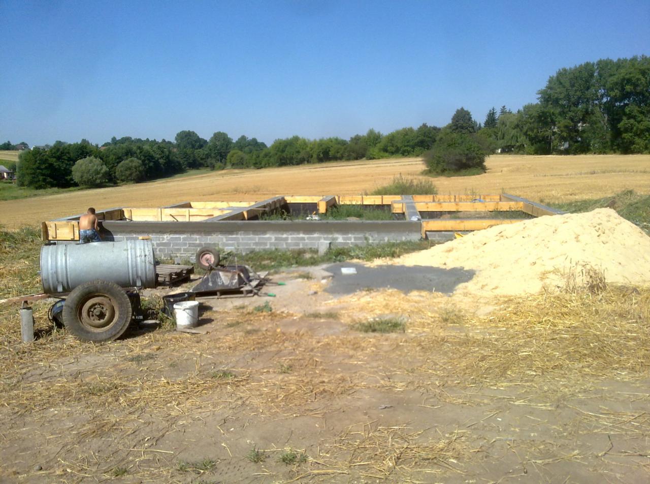Wylane fundamenty z szalunkiem na tle pola i lasu, widoczna betoniarka i sterta piasku, pracownik siedzący na betoniarce.
