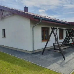 Wykończony dom jednorodzinny z jasną elewacją, brązową dachówką i drewnianą konstrukcją pod dachem, widok z boku na tle błękitnego nieba z chmurami, obok domu widoczna drewniana huśtawka.