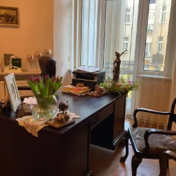 Biuro z ciemnym drewnianym biurkiem, na którym stoi wazon z fioletowymi tulipanami, globus, ramka ze zdjęciem, oraz statuetka Temidy w tle przy oknie.