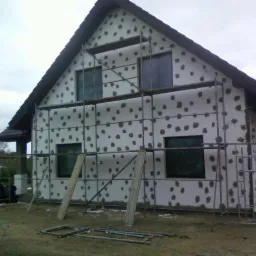 Dom jednorodzinny w trakcie ocieplania, widoczne rusztowania i płyty styropianowe z charakterystycznymi otworami na klej, przygotowane do dalszych prac wykończeniowych.