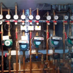 Rozbudowana instalacja hydrauliczna z manometrami i pompami obiegowymi Wilo, widoczne liczne rury miedziane i zawory z czerwonymi uchwytami, całość zamontowana na ścianie z folią izolacyjną.