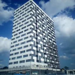 Wysoki budynek mieszkalny z elewacją w biało-szare pasy, częściowo pokryty rusztowaniami, na tle błękitnego nieba z chmurami.