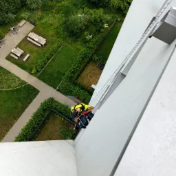 Pracownik w uprzęży, widok z góry podczas pracy na wysokości na białej elewacji budynku z widokiem na park z ławkami i zielonym dachem