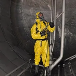 Pracownik w żółtym kombinezonie ochronnym i masce, spryskuje wnętrze metalowego zbiornika z widocznymi rurami.