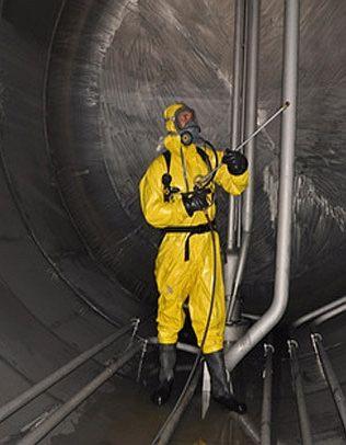 Pracownik w żółtym kombinezonie ochronnym i masce, spryskuje wnętrze metalowego zbiornika z widocznymi rurami.