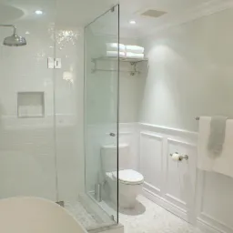 Jasna łazienka z kabiną prysznicową typu walk-in, chromowaną armaturą, białą ceramiką i mozaikową podłogą, wykończona w klasycznym stylu z panelami ściennymi.