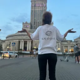 Osoba w białej koszulce z nadrukiem 'KAJ JO JEST' na tle Pałacu Kultury w Warszawie, z rozłożonymi rękami, prezentuje unikalny wzór koszulki.