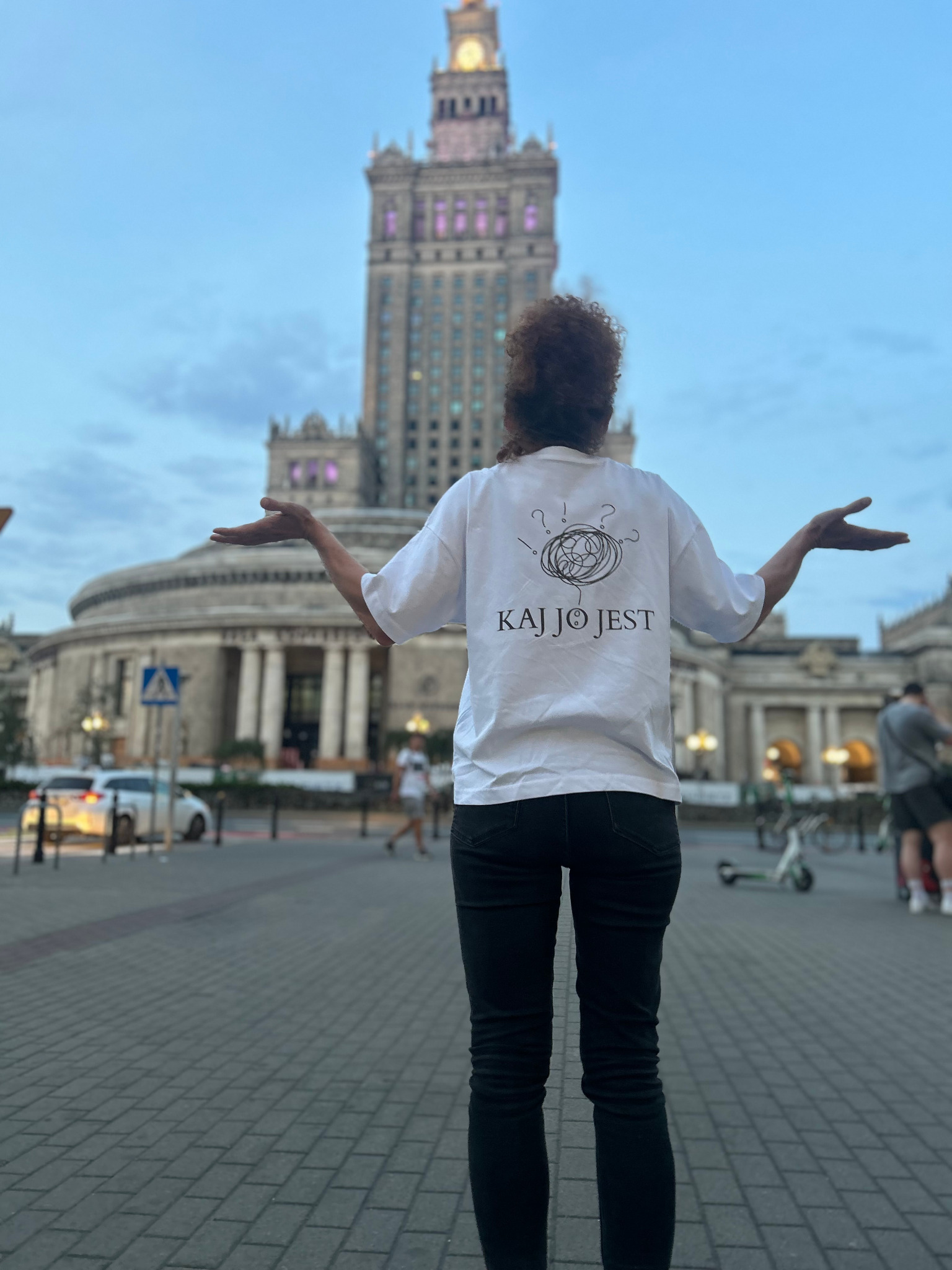 Osoba w białej koszulce z nadrukiem 'KAJ JO JEST' na tle Pałacu Kultury w Warszawie, z rozłożonymi rękami, prezentuje unikalny wzór koszulki.