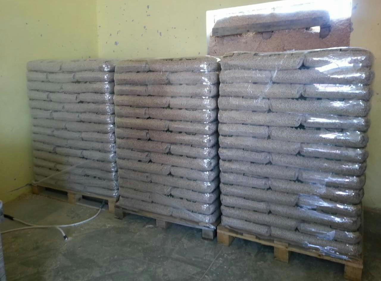 Trzy palety z workami pelletu, owinięte folią, ustawione w magazynie z widocznym oknem z drewnianą belką.