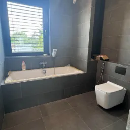 Wykończona łazienka w szarości. Nowoczesna toaleta, wanna zabezpieczona folią, okno z żaluzjami. Minimalistyczny design z ciemnymi płytkami i wiszącą lampą.