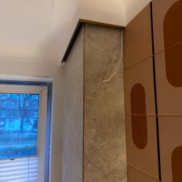 TK Format - Wykończony narożnik ściany z płytami imitującymi kamień, z metalową listwą. Obok ściana z dekoracyjnymi panelami w odcieniach brązu. W tle okno z żaluzjami.