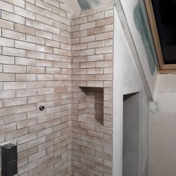 TK Format - Wykończona ściana z beżowych płytek w łazience na poddaszu, z wnęką i widoczną instalacją. Ściany przygotowane do dalszych prac wykończeniowych.