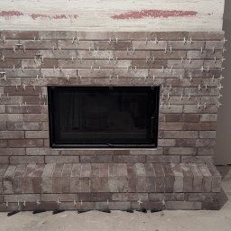 TK Format - Obudowa kominka z cegieł w trakcie montażu, z krzyżykami dystansowymi. Widoczne narzędzia i materiały budowlane. Ceglana faktura z czarnym wkładem kominkowym.