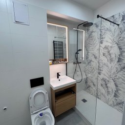 TK Format - Nowoczesna łazienka po remoncie: białe płytki, kabina prysznicowa z motywem liści, umywalka z szafką z jasnego drewna i podświetlane lustro. Minimalistyczny design.